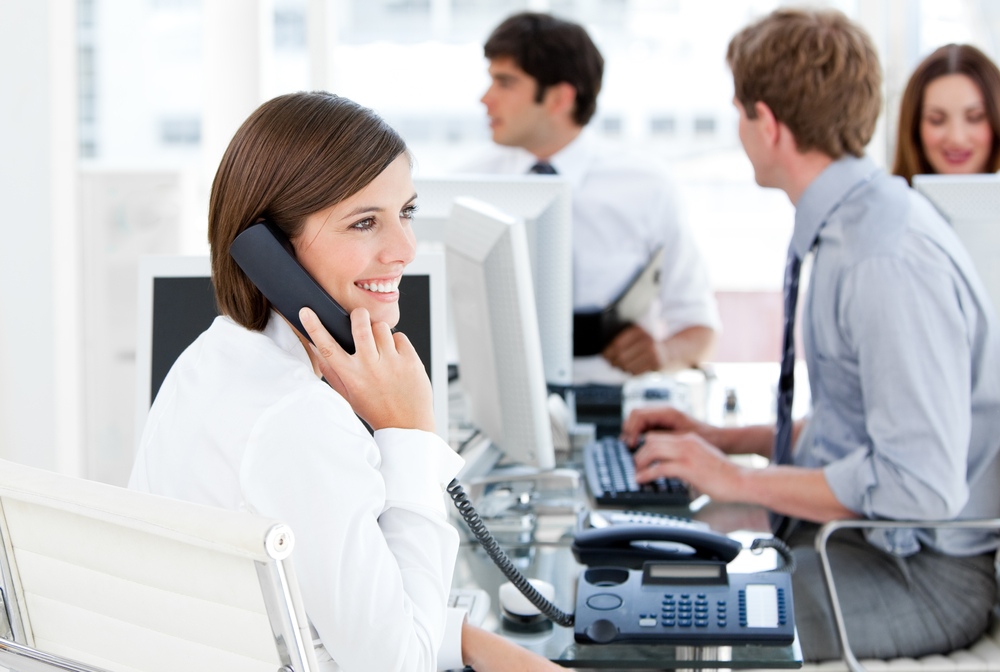 A stock image depicting a smiling woman answering an office phone while 3 colleagues work in the background