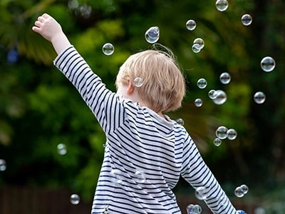 A young boy with blonde hair reaching up to pop soap bubbles.