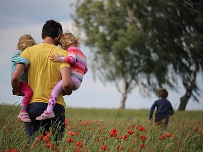 A father carrying his two young children on his hips through a field of red poppies.
